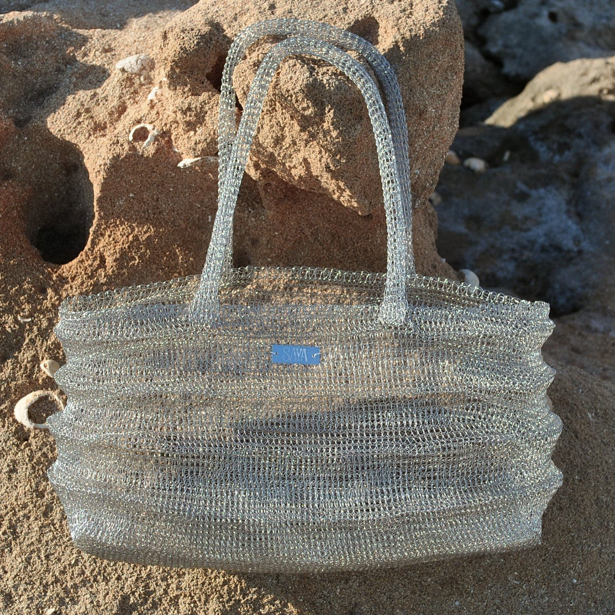 Bolso Ombak plata tejido a mano en crochet metálico sobre rocas junto al mar.