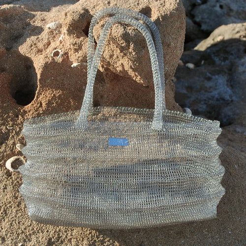 Bolso Ombak plata tejido a mano en crochet metálico sobre rocas junto al mar.